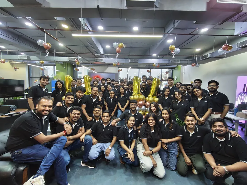 PromotEdge team members in black polo shirts, sitting and standing together in the office with gold balloons.